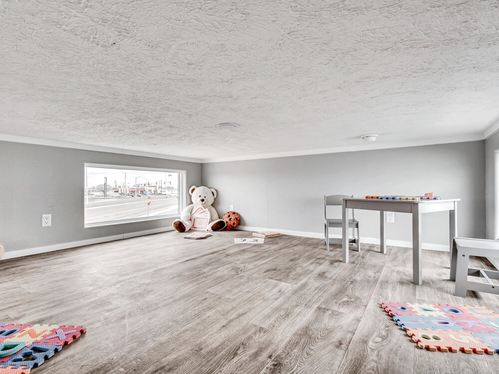 Children's playroom with a gray table, chairs, and colorful foam mats on wooden flooring. A plush teddy bear and toys sit in the corner near a large window.