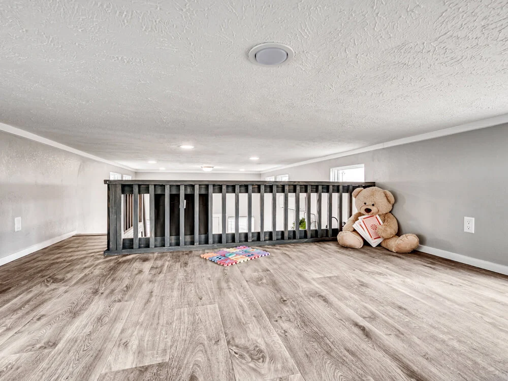 Loft area with light wood flooring and gray walls. A large teddy bear holds a book next to a colorful rug. The space feels cozy and playful.