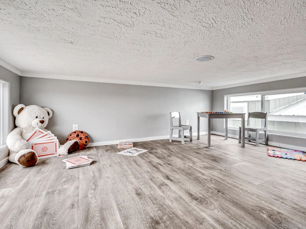 A cozy playroom with a low ceiling features a large stuffed bear holding oversized playing cards, a ladybug toy, and a small table with chairs by a window.