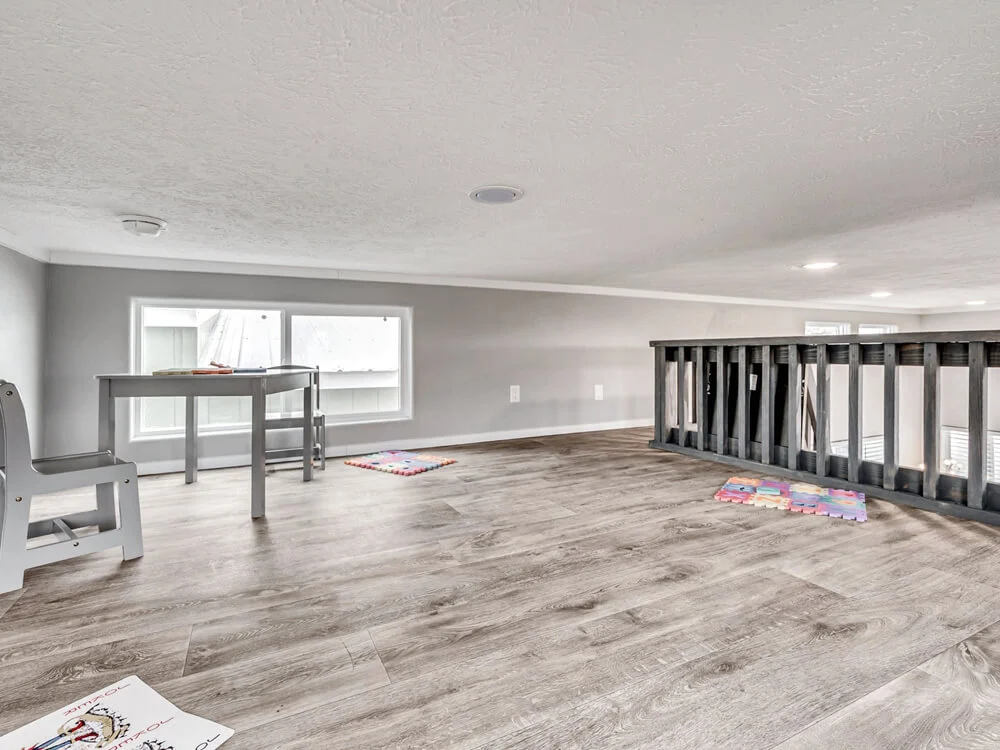 Loft play area with gray walls and wooden floor. Features a small table and chair set near a window, colorful foam mats, and a railing overlooking below.