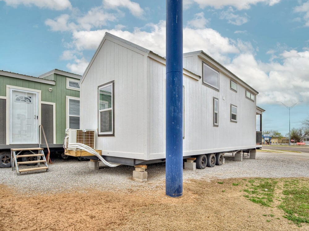 A white mobile tiny home on wheels is parked on a gravel surface next to a green tiny home. A large blue pole stands in the foreground. The sky is partly cloudy.