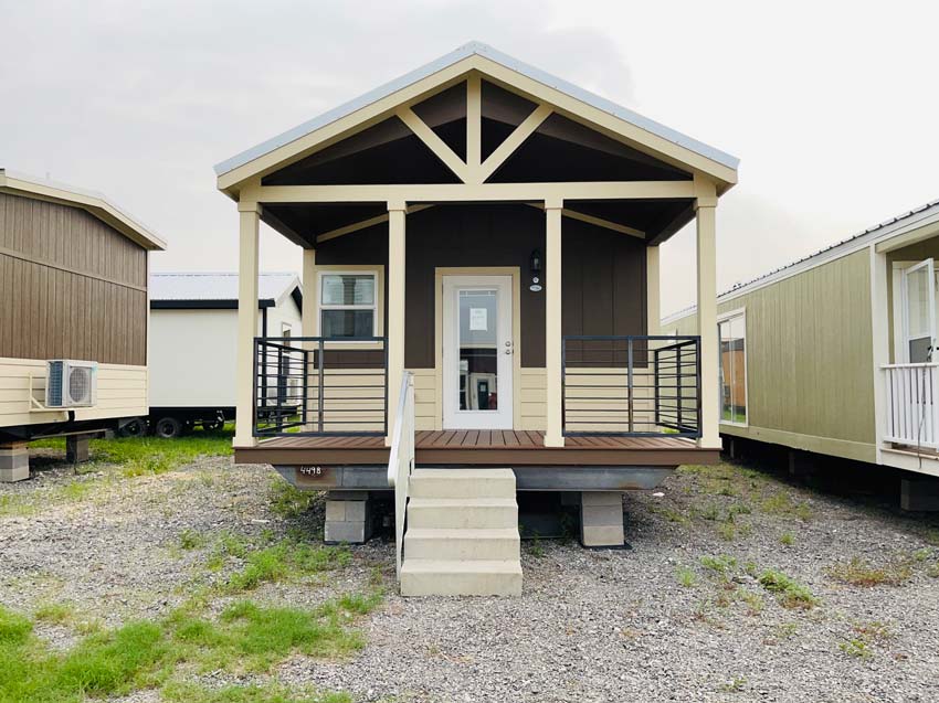 Small, modern prefab house with a brown and beige exterior, front porch, and steps. It's on a gravel lot, surrounded by similar structures.