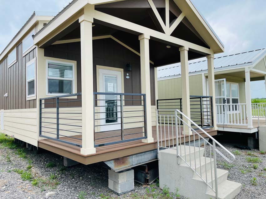 Small brown and cream modular home with a covered porch and metal railing. Concrete steps lead up to a glass door. Overcast sky in the background.