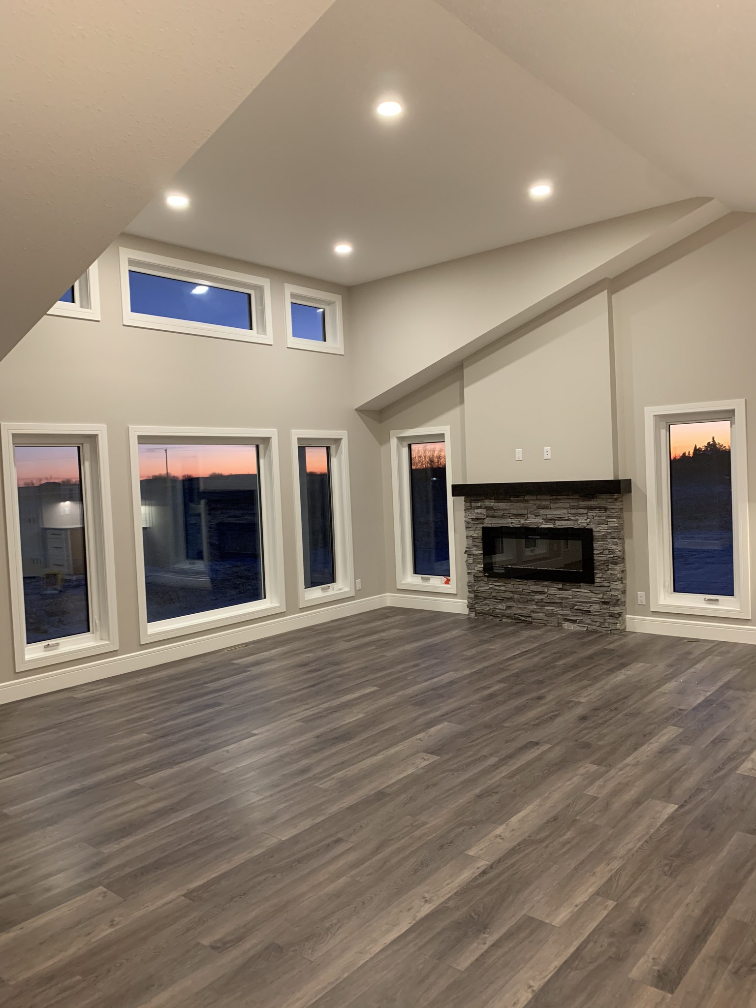 Spacious living room with large windows, sunset view, gray walls, vaulted ceiling, recessed lighting, stone fireplace, and dark wood flooring.