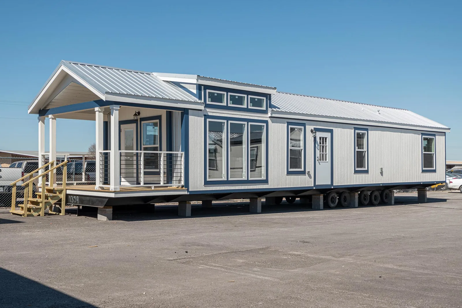 A modern tiny house on wheels with a metal roof and large windows stands in a sunny lot. It features a small porch with railing, evoking a cozy and inviting vibe.