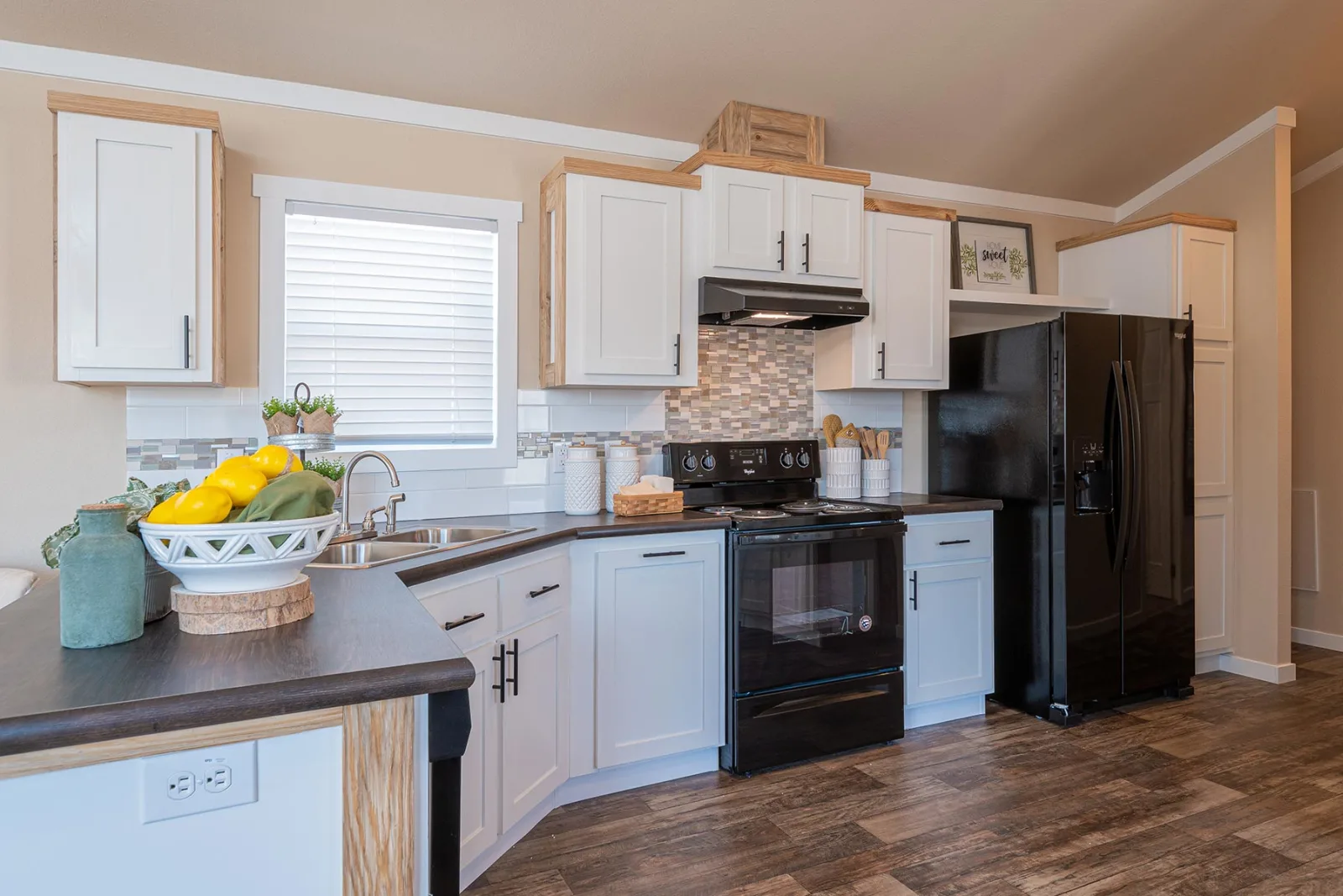 Modern kitchen with white cabinets, black appliances, and dark countertops. Bowl of lemons, plants, and wooden utensils add a fresh and cozy touch.