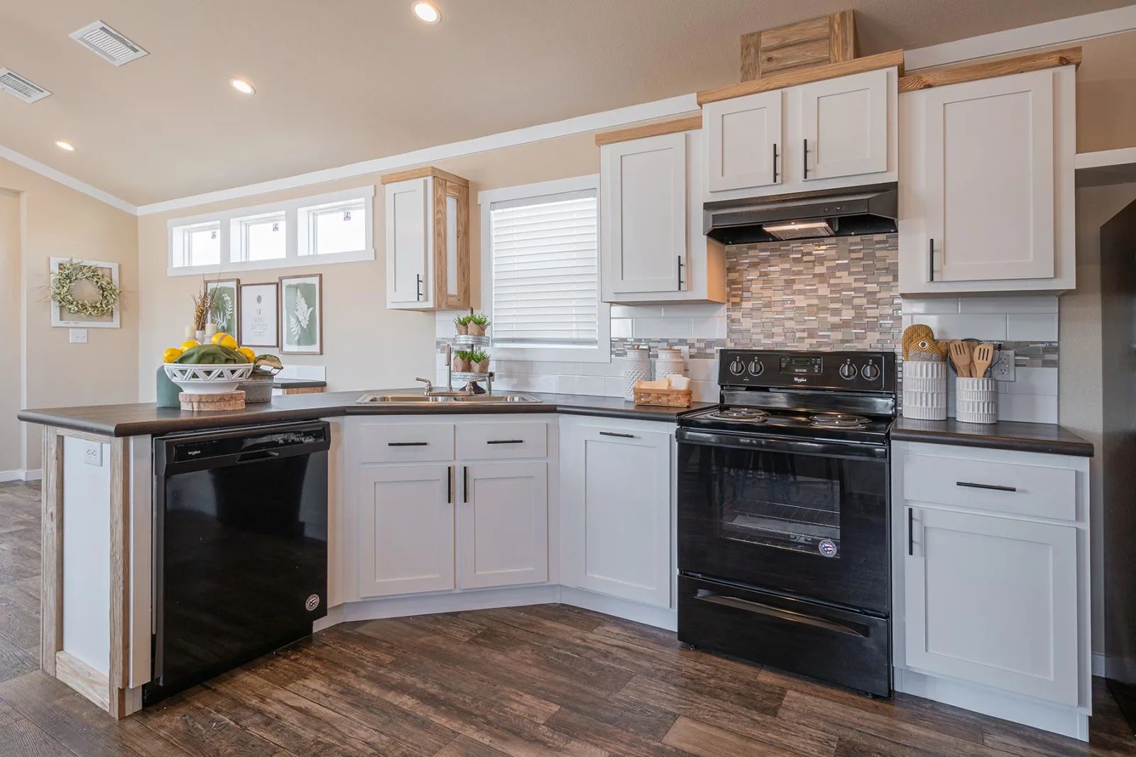 Modern kitchen with white cabinets, black appliances, and a mosaic tile backsplash. A countertop holds a fruit basket, and wood floors add warmth.