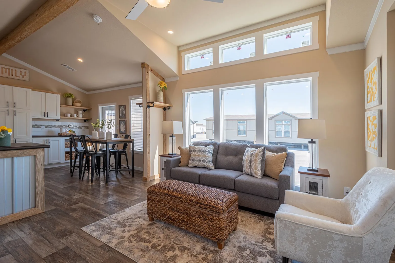Bright, inviting living room with large windows, gray sofa, patterned cushions, wicker ottoman, and a cozy armchair. Open kitchen and dining area. Warm tones.