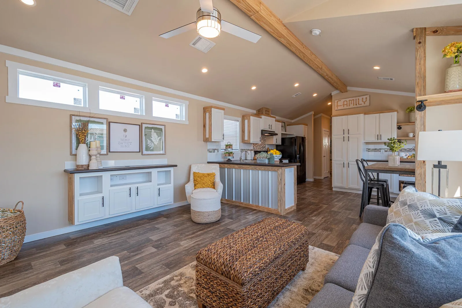 Modern open-plan living area with a cozy vibe. Features a kitchen island, wicker ottoman, wooden beams, and neutral tones. Natural light brightens the space.