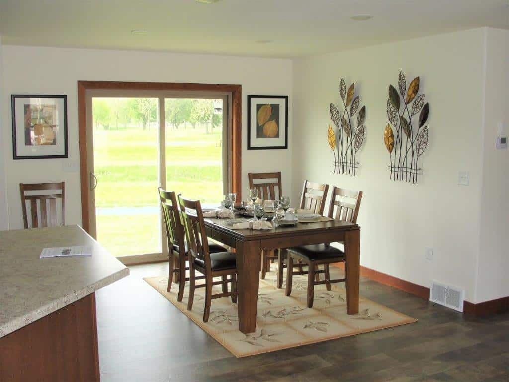 A cozy dining room with a wooden table set for six, adorned with glassware and napkins. Sliding doors reveal a lush green view, creating a serene ambiance. Leaf-themed wall art and framed prints add a natural touch.
