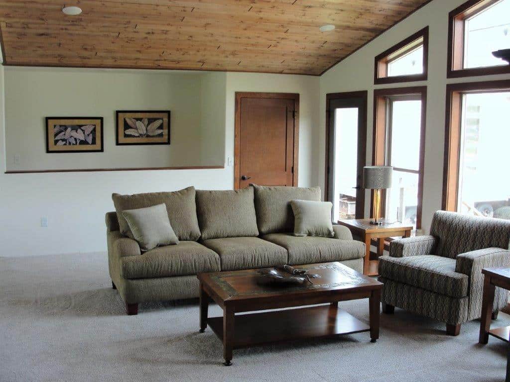 A cozy living room with beige carpet features a taupe sofa, patterned armchair, and wooden coffee table. Soft light from large windows creates a warm, inviting atmosphere. Framed artwork hangs on the wall, and a wooden door adds a rustic touch.