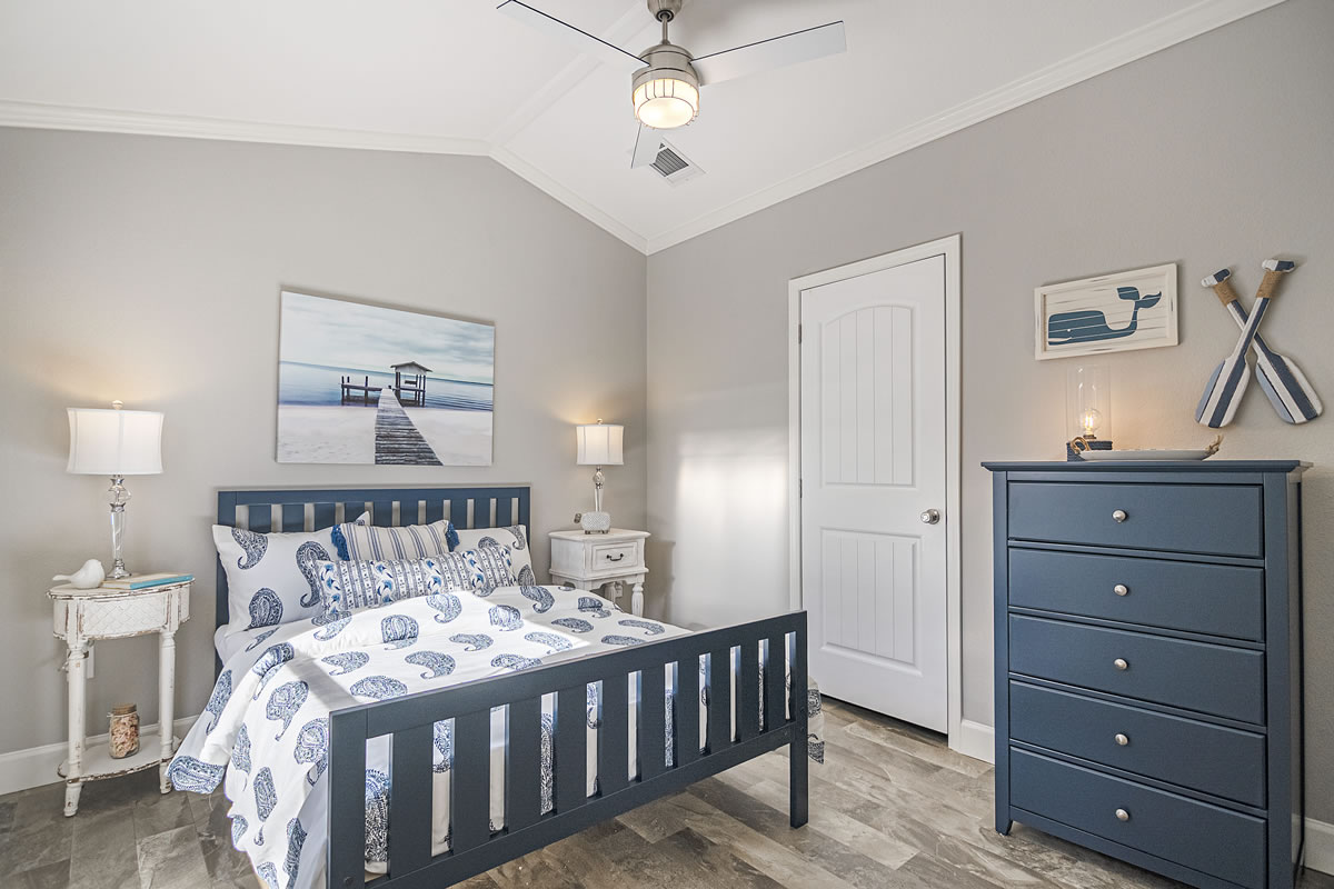 A cozy bedroom features a nautical theme with a blue bed and dresser. Ocean artwork and decorative oars adorn gray walls, creating a serene, beach-like ambiance.