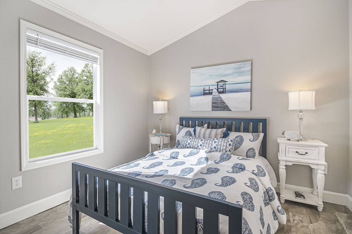 A cozy bedroom with a dark blue wooden bed, covered in a white and blue paisley-patterned comforter. Two white nightstands with lamps flank the bed, beneath a serene ocean pier painting. A window on the left shows a sunny green landscape. The room exudes a calm and inviting atmosphere.