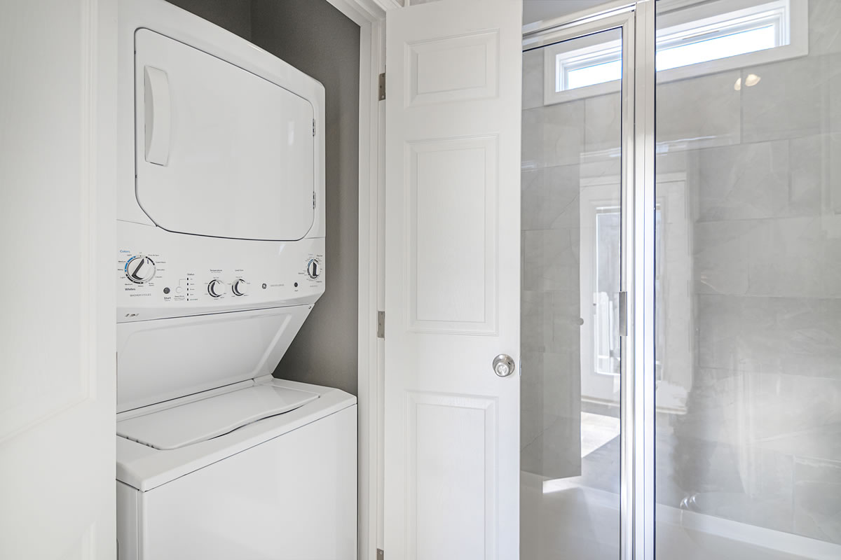 Stacked white washer and dryer in a small laundry closet with an open white door. Sunlit bathroom with glass shower enclosure is visible nearby.