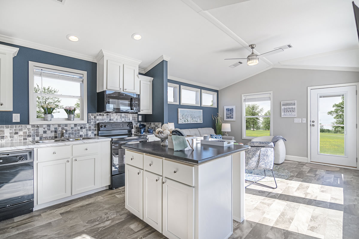 Modern kitchen and living area with white cabinets, black appliances, and a central island. Large windows reveal a green landscape outside. Airy and bright ambiance.