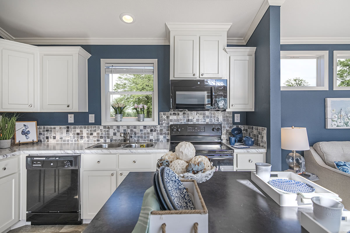 Modern kitchen with white cabinets, blue walls, and mosaic backsplash. A window above the sink reveals greenery outside. Cozy atmosphere with a basket on a counter and decorative items.