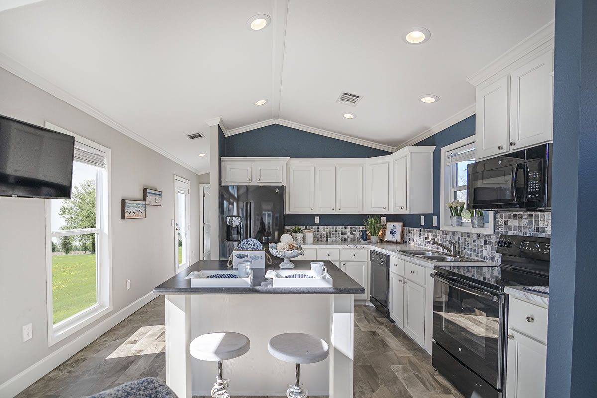Modern kitchen with white cabinets, black appliances, and a central island with two stools. Blue-gray walls, a patterned backsplash, and large window enhance brightness.