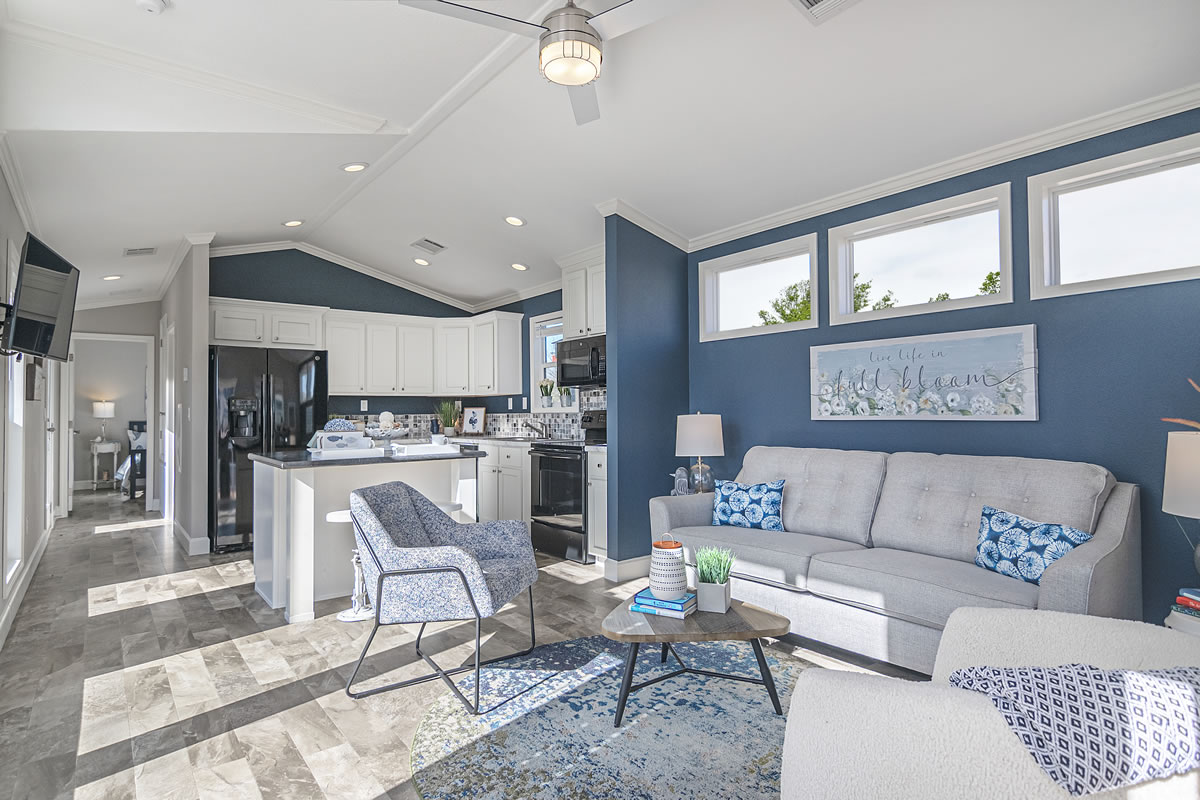 Bright living room with blue accents featuring a gray couch, matching chairs, a patterned rug, and a coffee table. A kitchen with white cabinets and a dark island is visible, enhancing the modern feel.