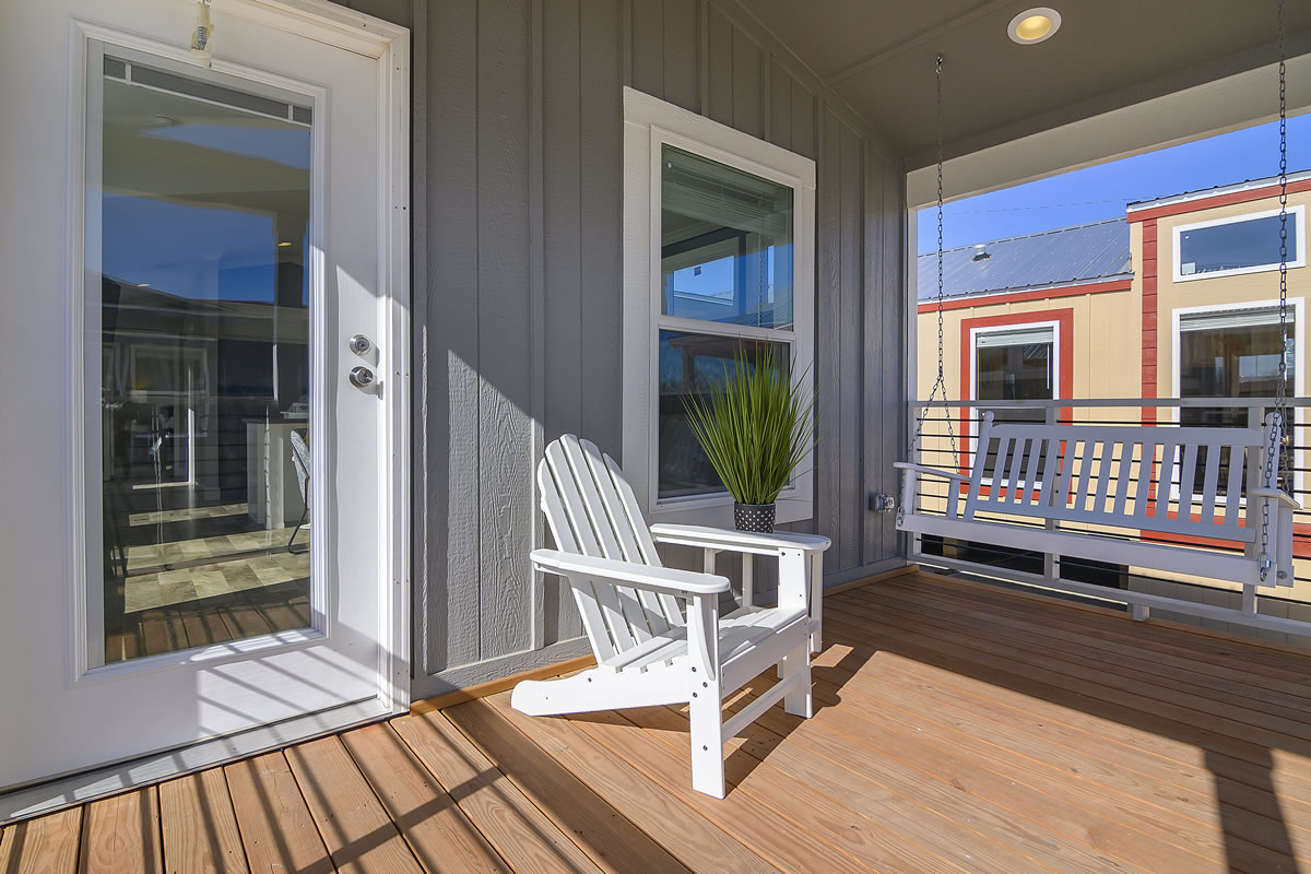 Outdoor porch with a wooden floor, featuring a white Adirondack chair and a matching swing. A potted plant sits between them against a gray wall.