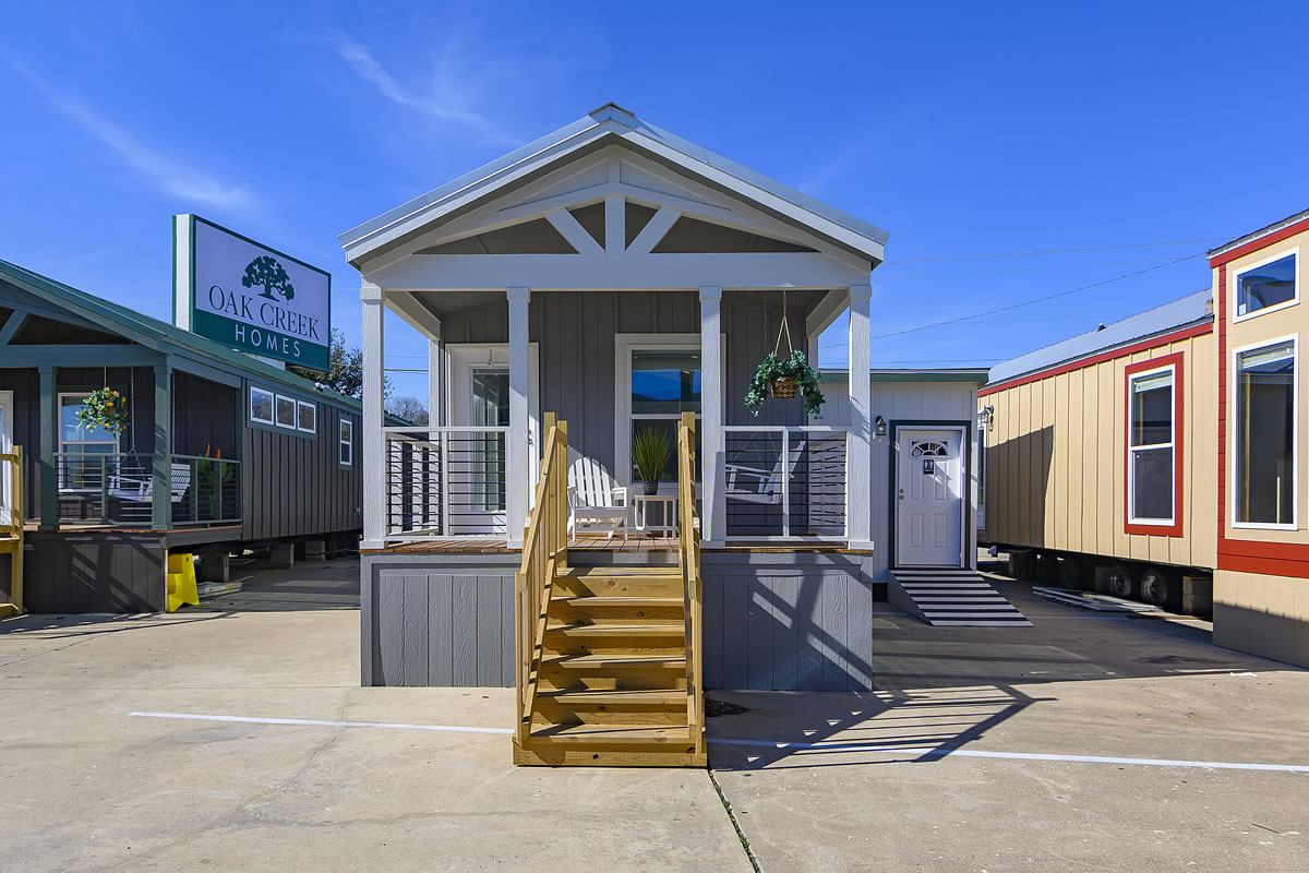Tiny house model under a clear blue sky, featuring a front porch with wooden steps, hanging plants, and a sign for Oak Creek Homes in the background.