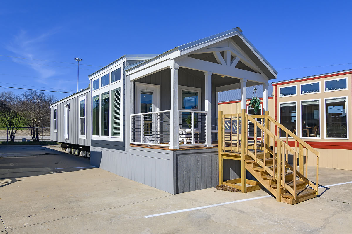 Modern gray tiny house with a gabled roof and large windows sits on a concrete lot. Wooden steps lead to a porch, under a clear blue sky.