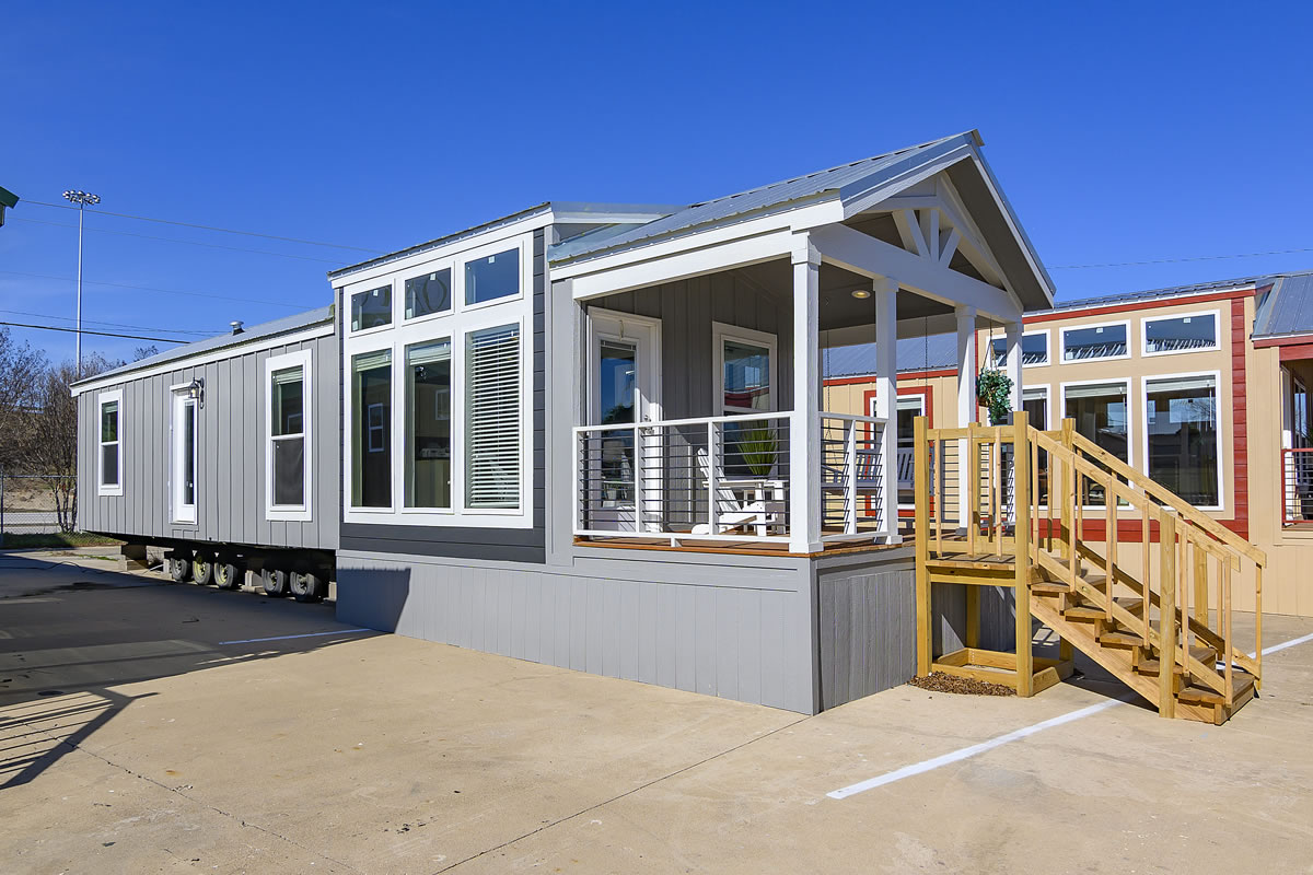 Modern mobile home with large windows and a small porch sits on a concrete lot under a clear blue sky. Wooden stairs lead to the entrance.