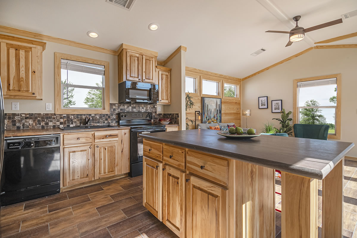 Spacious kitchen with wooden cabinets, tiled backsplash, and dark countertops. Sunlit living area with large windows, plants, and a ceiling fan. Cozy, natural ambiance.