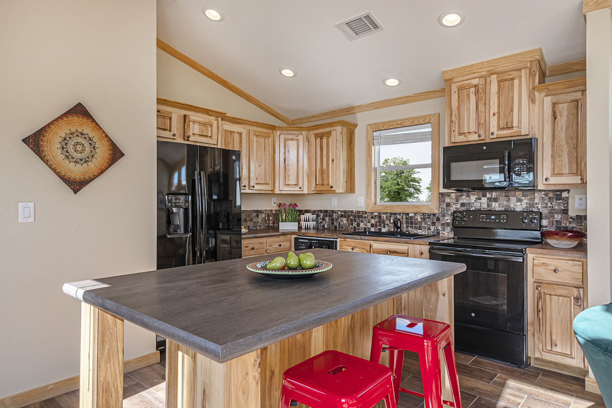 Warm, inviting kitchen with natural wood cabinets, tiled backsplash, black appliances, and a central island. Red stools and green apples add vibrant pops.
