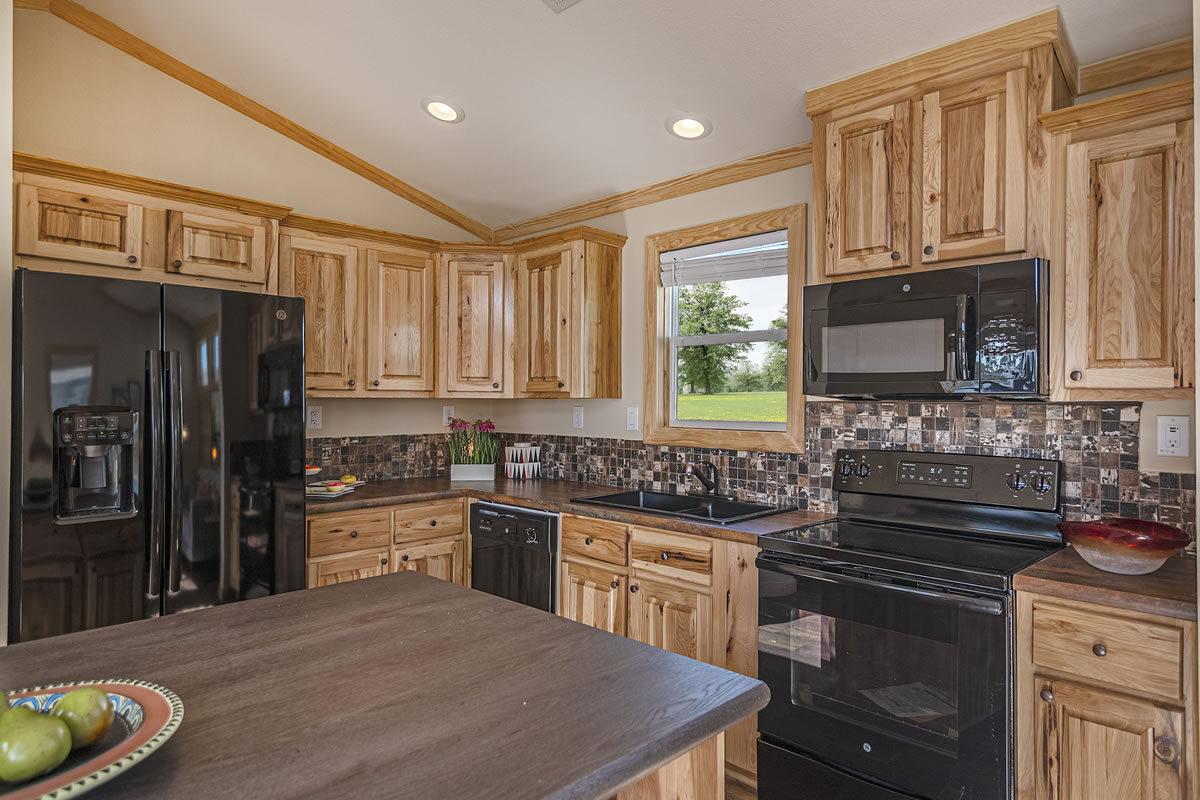 Rustic kitchen with wooden cabinets, black appliances, and a dark island. Mosaic backsplash adds texture. Window shows a green field outside.