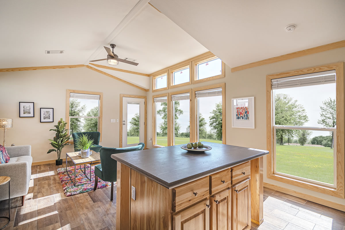 Bright, modern living room with a large island in the foreground. Sunlight streams through tall windows, highlighting green trees and a cozy interior with colorful accents.