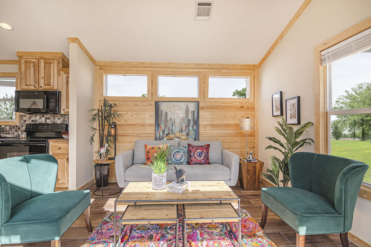 Bright living room with wooden paneling, a gray sofa with colorful pillows, and green armchairs. A coffee table, plants, and abstract art adorn the space.