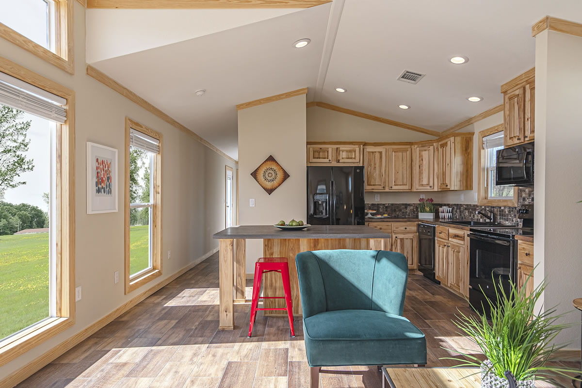 Bright, modern kitchen with wood cabinets, a black fridge, and a center island. A red stool, teal chair, and lush greenery add vibrant accents.