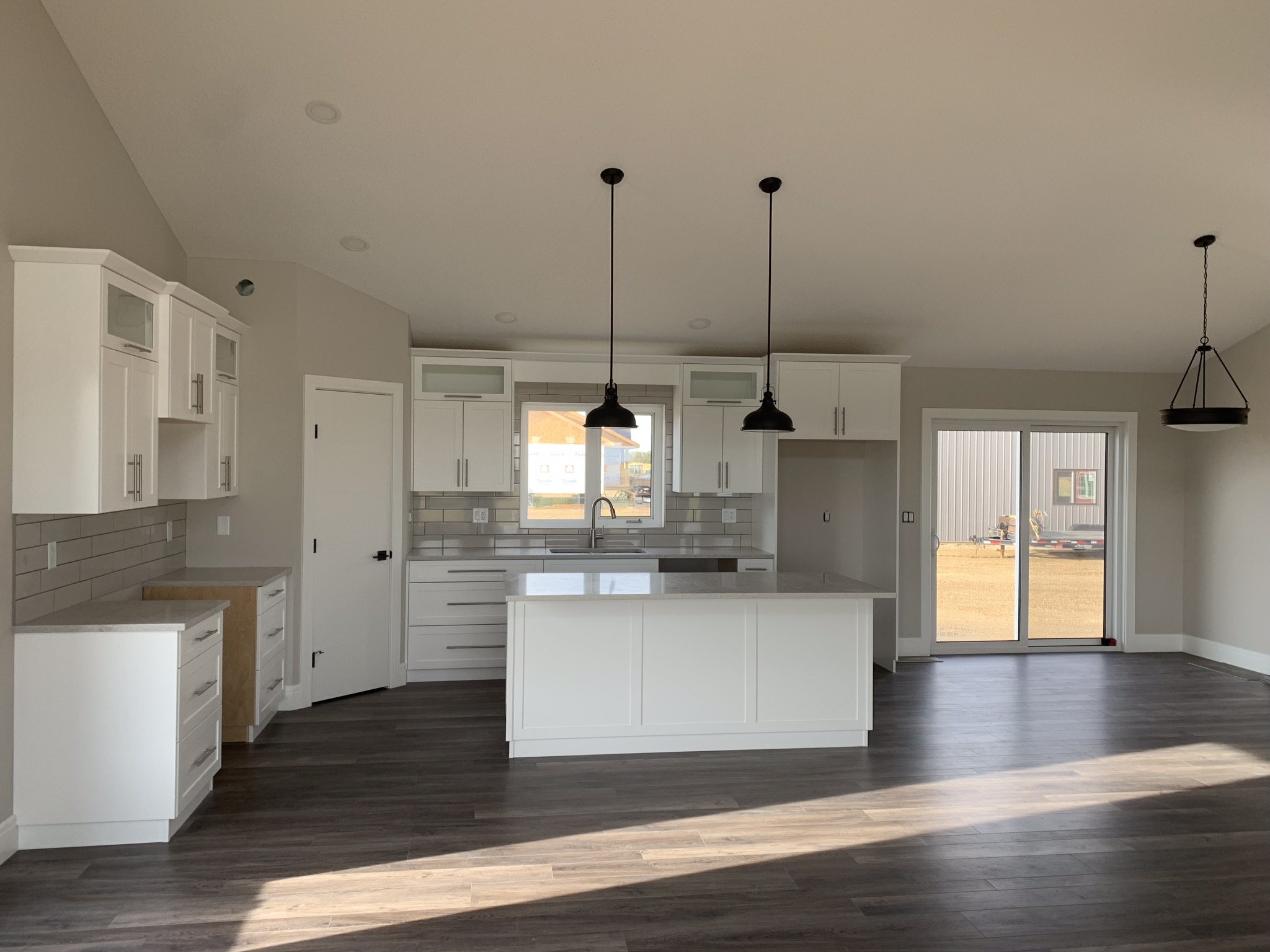 Spacious kitchen with white cabinets, dark wood floor, and an island. Black pendant lights hang above. Sunlight streams through glass doors. Minimalist and modern.