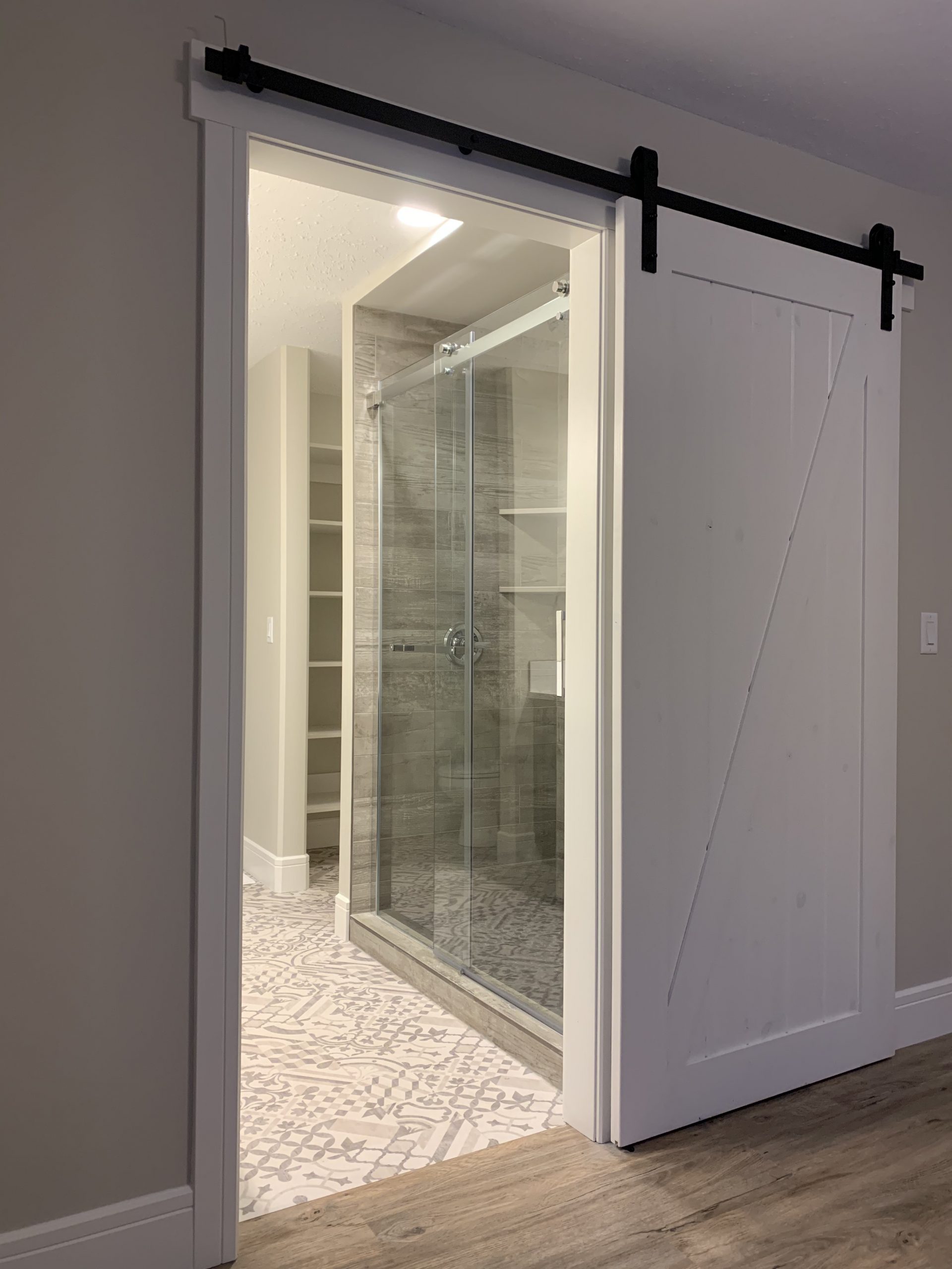 A modern bathroom with a glass shower is visible through a partially open white barn-style sliding door. The room has patterned tiles and a sleek design.
