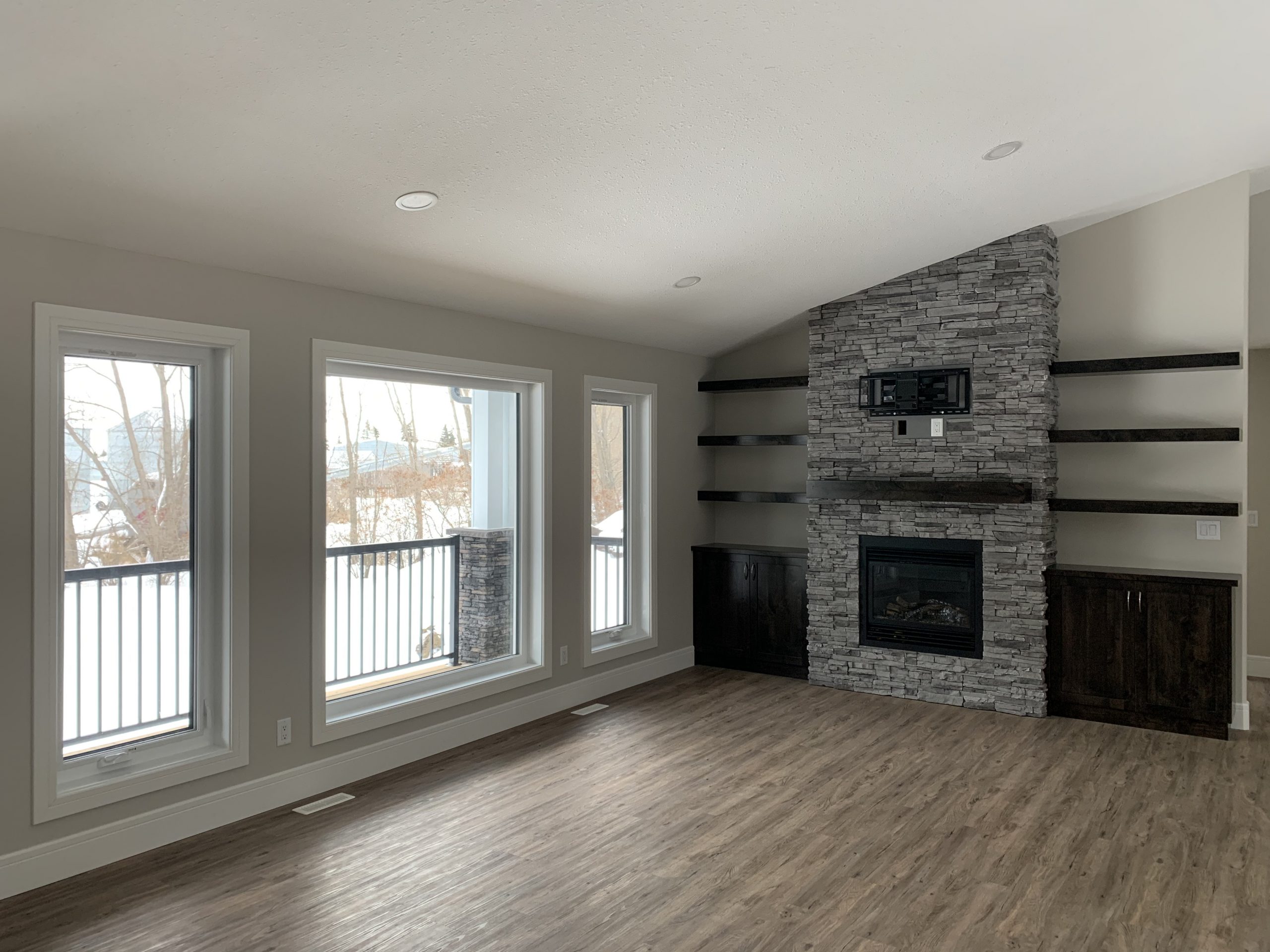 Modern living room with large windows, wood flooring, and a stone fireplace. Dark wooden shelves flank the fireplace, and snow is visible outside.