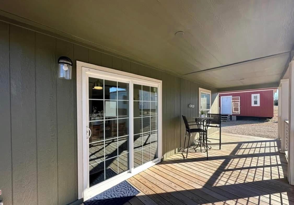 Covered wooden porch with a small table and chairs. Sliding glass doors lead inside, and a red outbuilding is visible on gravel in the background. Sunny atmosphere.