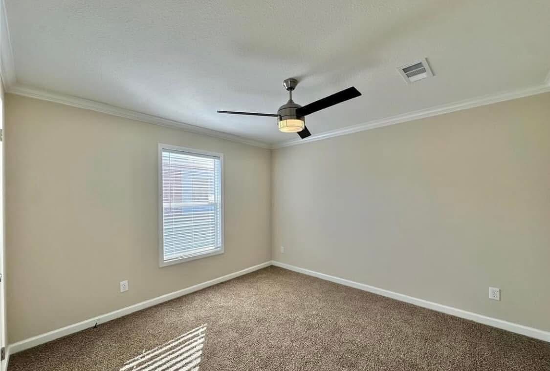 An empty room with beige walls and carpet, featuring a large window with blinds and a modern black ceiling fan. The space feels bright and minimalist.