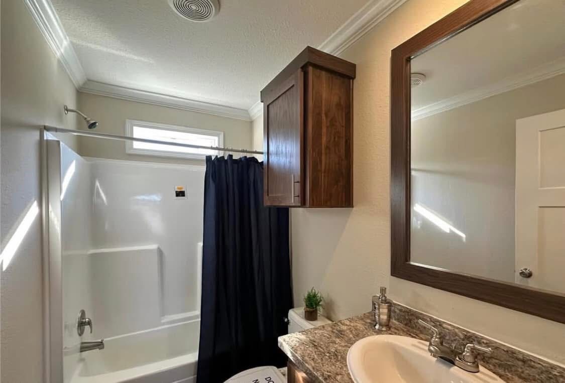 A small bathroom with a dark shower curtain, white tub, and wooden cabinets. The sink has a granite countertop, and the space is lit by a small window.