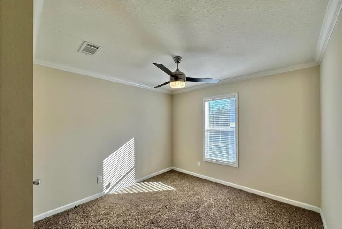 A small, empty room with beige walls and brown carpet. A ceiling fan with a light fixture is centered above. Sunlight filters through a window with blinds, creating patterns on the wall, conveying a calm and peaceful atmosphere.