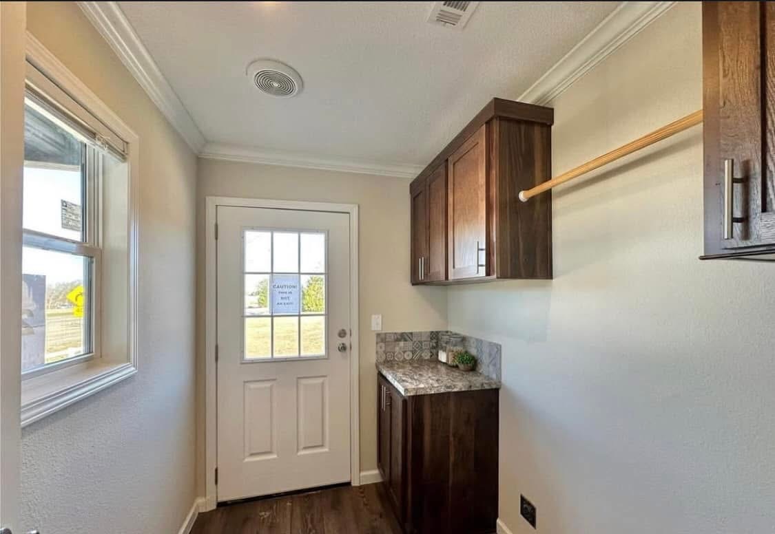 Narrow laundry room with a windowed door, sunlight streaming in. Dark wooden cabinets, marble counter, and a wooden rod for hanging clothes.