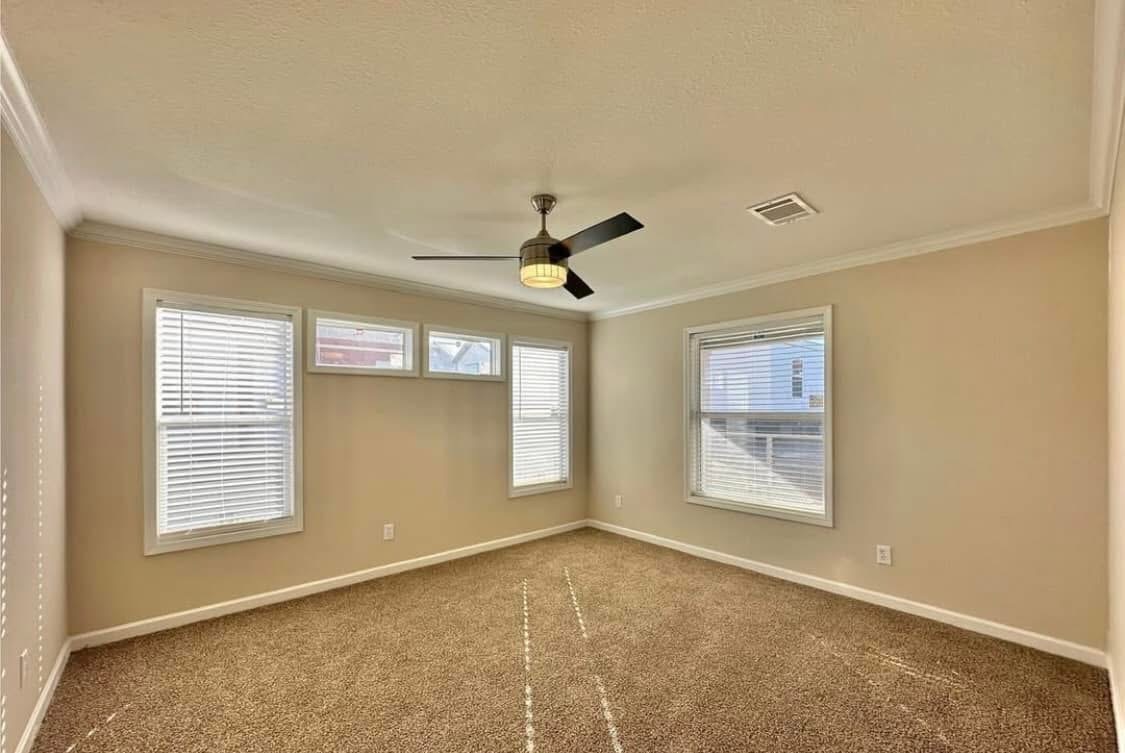 A spacious, empty room with beige walls and carpeted floors. Four windows allow natural light. A modern ceiling fan hangs in the center, creating an airy vibe.