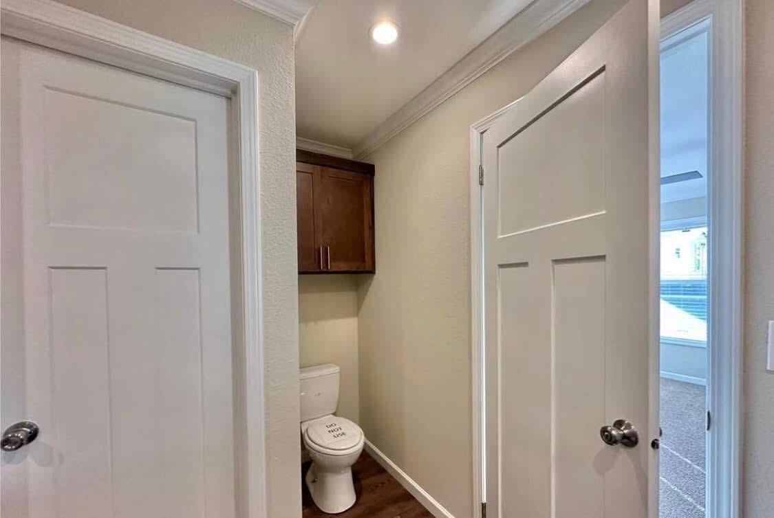 A small, well-lit bathroom with beige walls, featuring a toilet with a humorous message. Dark wood cabinet above, white doors, and hardwood floor.