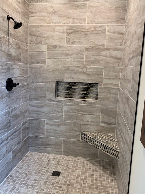 Modern shower with gray tile walls and floor, featuring a small black showerhead, built-in niche, and corner bench with contrasting mosaic tiles.