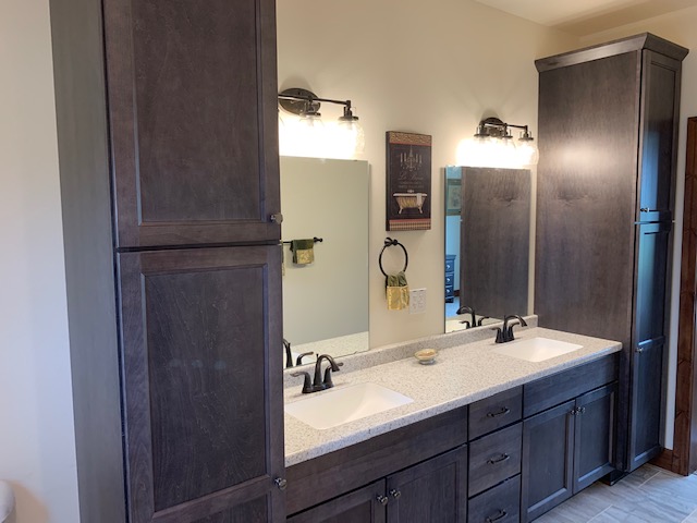 Modern bathroom with dark wood cabinets and granite countertop. Two sinks with black faucets are below lit mirrors. Walls are light-colored.