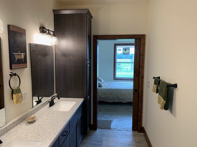 Modern bathroom with double sinks and dark cabinets on the left, light gray counters, and a warmly-lit mirror. A doorway leads to a bedroom.