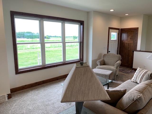 Bright living room with large windows showing a green field. Beige furniture, a lamp, and wooden door create a cozy, inviting atmosphere.