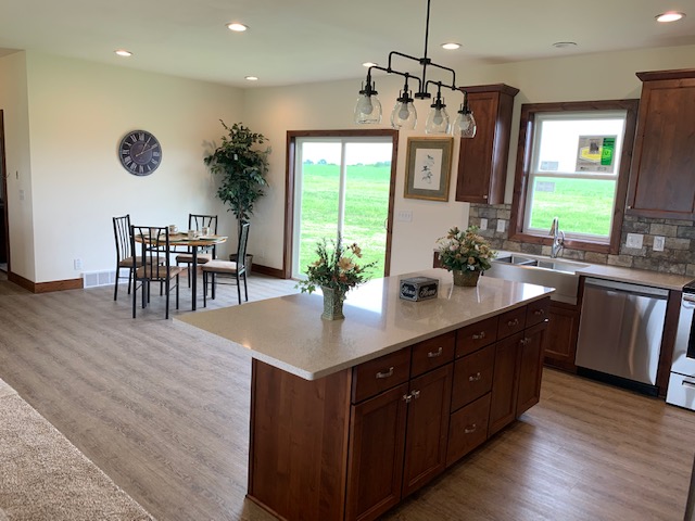 Modern kitchen with wooden cabinets and a central island featuring floral decor. A dining table, large windows, and warm lighting create a cozy ambiance.