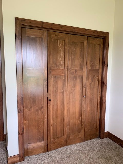 Wooden closet doors with a natural finish in a carpeted room. The doors have a paneled design and small round knobs, creating a warm, rustic tone.