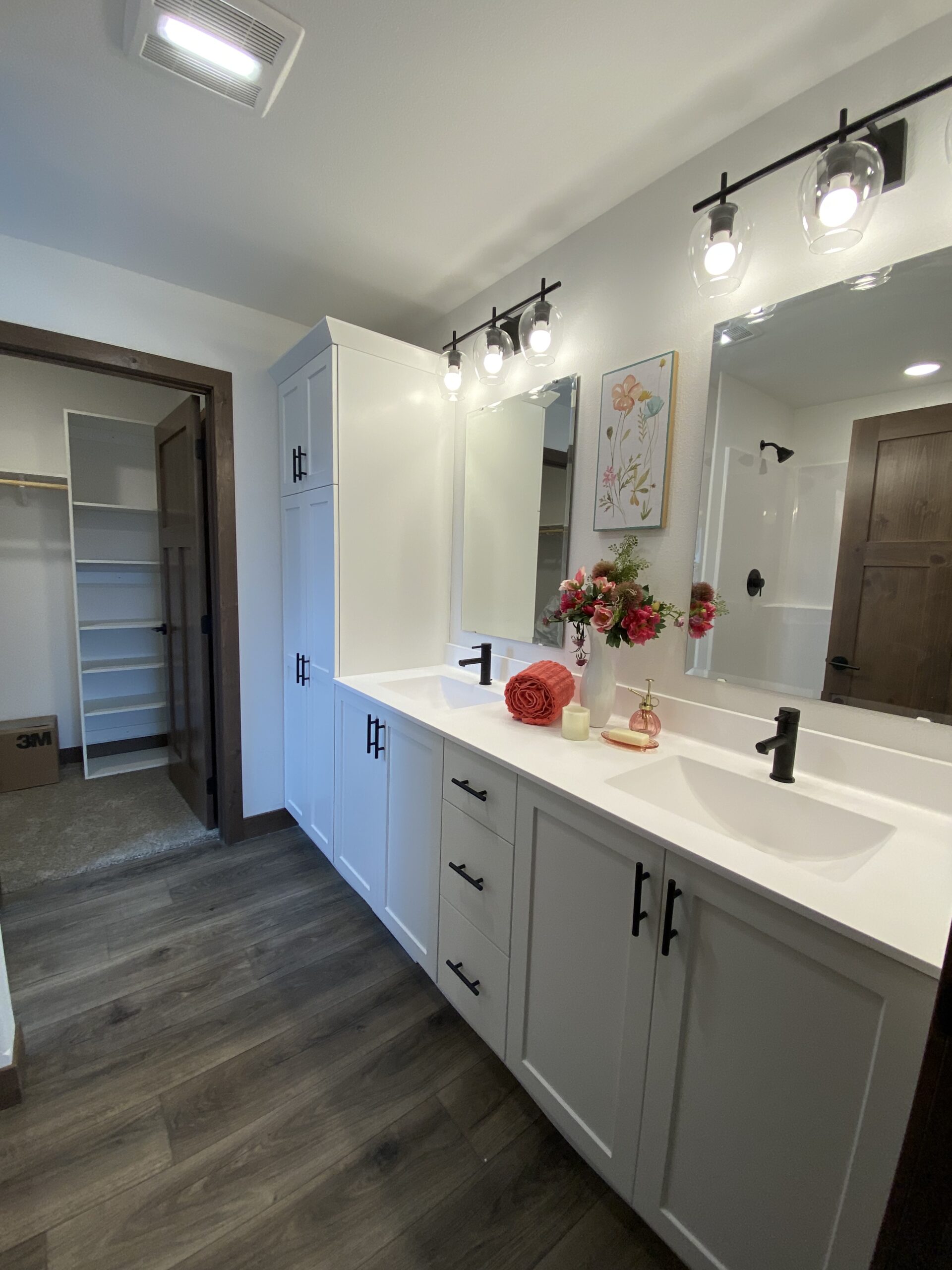 Modern bathroom with a white double-sink vanity, black fixtures, and wood floor. Soft lighting, floral decor, and a walk-in closet create a cozy ambiance.