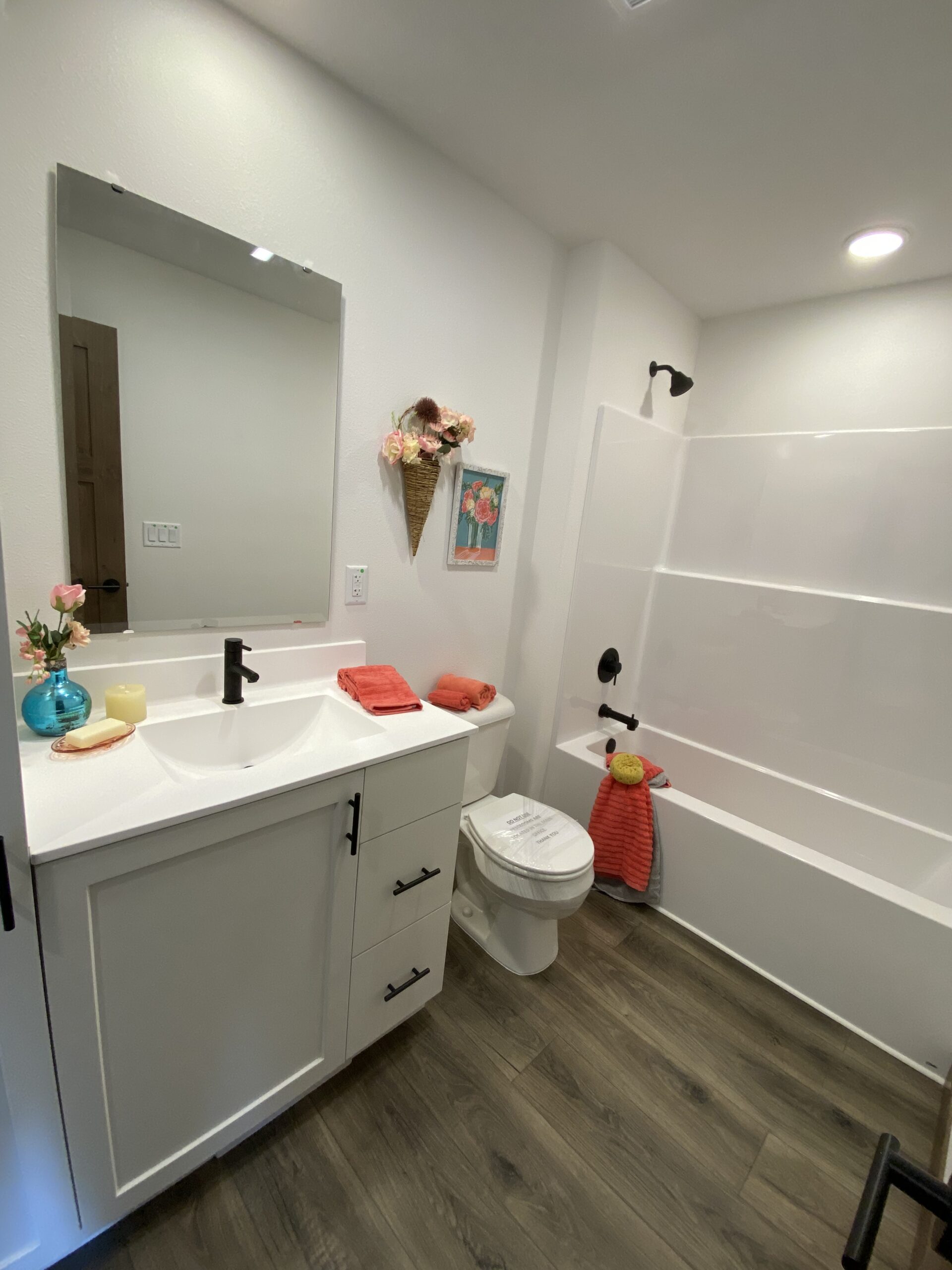 Modern bathroom with white vanity and dark faucet, framed by vibrant red towels, and floral decor. White bathtub and wood floor add warmth.